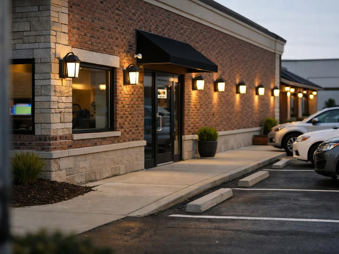 A brick storefront with lit wall lamps, a glass door, and parked cars lines a quiet sidewalk at dusk